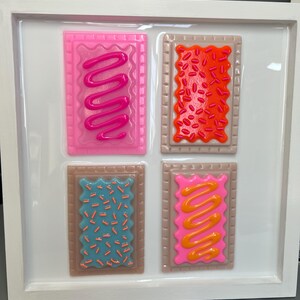May include: A framed artwork featuring four colorful, stylized Pop-Tart-like designs. Each rectangular piece has a brown border and a different frosting and sprinkle design in pink, orange, blue, and yellow. The artwork is set against a white background.