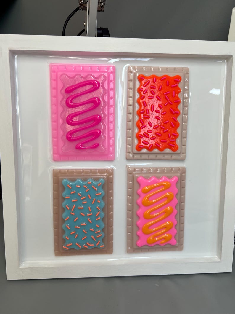 May include: A framed artwork featuring four colorful, stylized Pop-Tart-like designs. Each rectangular piece has a brown border and a different frosting and sprinkle design in pink, orange, blue, and yellow. The artwork is set against a white background.