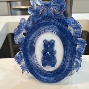 Op de afbeelding: Een blauw en wit keramiek fotolijstje met een blauwe glitterafwerking. Het lijstje heeft een decoratieve strik en een wit ovaal met een blauwe gummybeer erin.