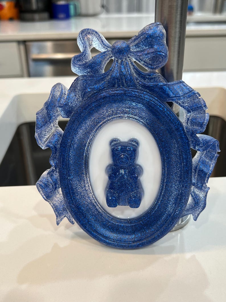 Op de afbeelding: Een blauw en wit keramiek fotolijstje met een blauwe glitterafwerking. Het lijstje heeft een decoratieve strik en een wit ovaal met een blauwe gummybeer erin.