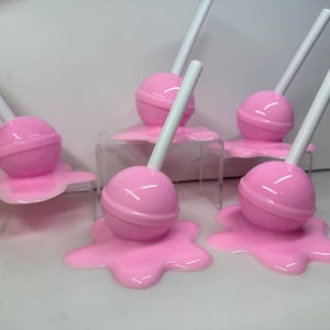 Op de afbeelding: Een verzameling roze lolly's met witte stokjes, die lijken te smelten op een roze, onregelmatige basis. De lolly's worden gepresenteerd op kleine, transparante standaards tegen een witte achtergrond.