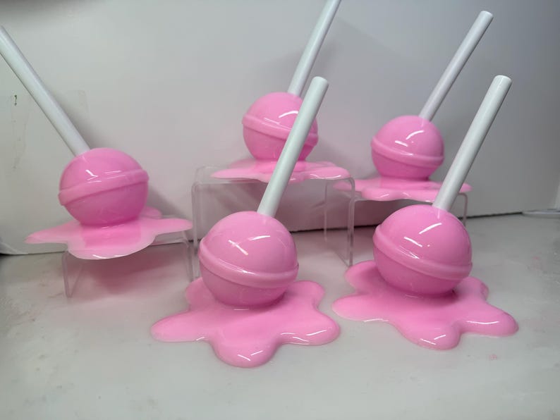 Op de afbeelding: Een verzameling roze lolly's met witte stokjes, die lijken te smelten op een roze, onregelmatige basis. De lolly's worden gepresenteerd op kleine, transparante standaards tegen een witte achtergrond.