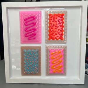 May include: A framed artwork featuring four colorful, rectangular pieces resembling frosted pastries. Each piece has a different color frosting and sprinkles, with a white frame. The pastries are pink, orange, blue, and pink with different frosting designs.