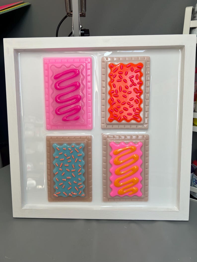 May include: A framed artwork featuring four colorful, rectangular pieces resembling frosted pastries. Each piece has a different color frosting and sprinkles, with a white frame. The pastries are pink, orange, blue, and pink with different frosting designs.