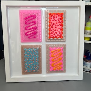 May include: A framed artwork featuring four colorful, stylized Pop-Tarts. Each pastry has a different frosting and sprinkle design: pink with pink frosting, orange with red sprinkles, blue with blue sprinkles, and pink with orange frosting. The frame is white.