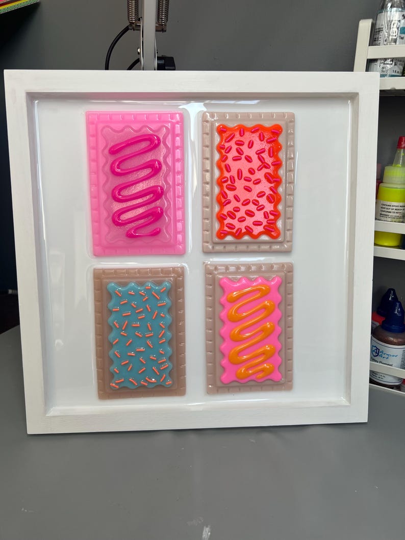 May include: A framed artwork featuring four colorful, stylized Pop-Tarts. Each pastry has a different frosting and sprinkle design: pink with pink frosting, orange with red sprinkles, blue with blue sprinkles, and pink with orange frosting. The frame is white.