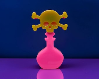 May include: A bright neon pink bottle-shaped object topped with a translucent yellow skull and crossbones emblem. The piece features a smooth, glossy finish and a distinct silhouette, with the skull element centred directly above the bottle neck.