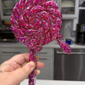 May include: A lollipop-shaped object featuring a swirling, circular design with a thick handle. The entire piece is crafted from a translucent, deep pink resin material embedded with a dense mixture of reflective, multi-sized pink glitter.