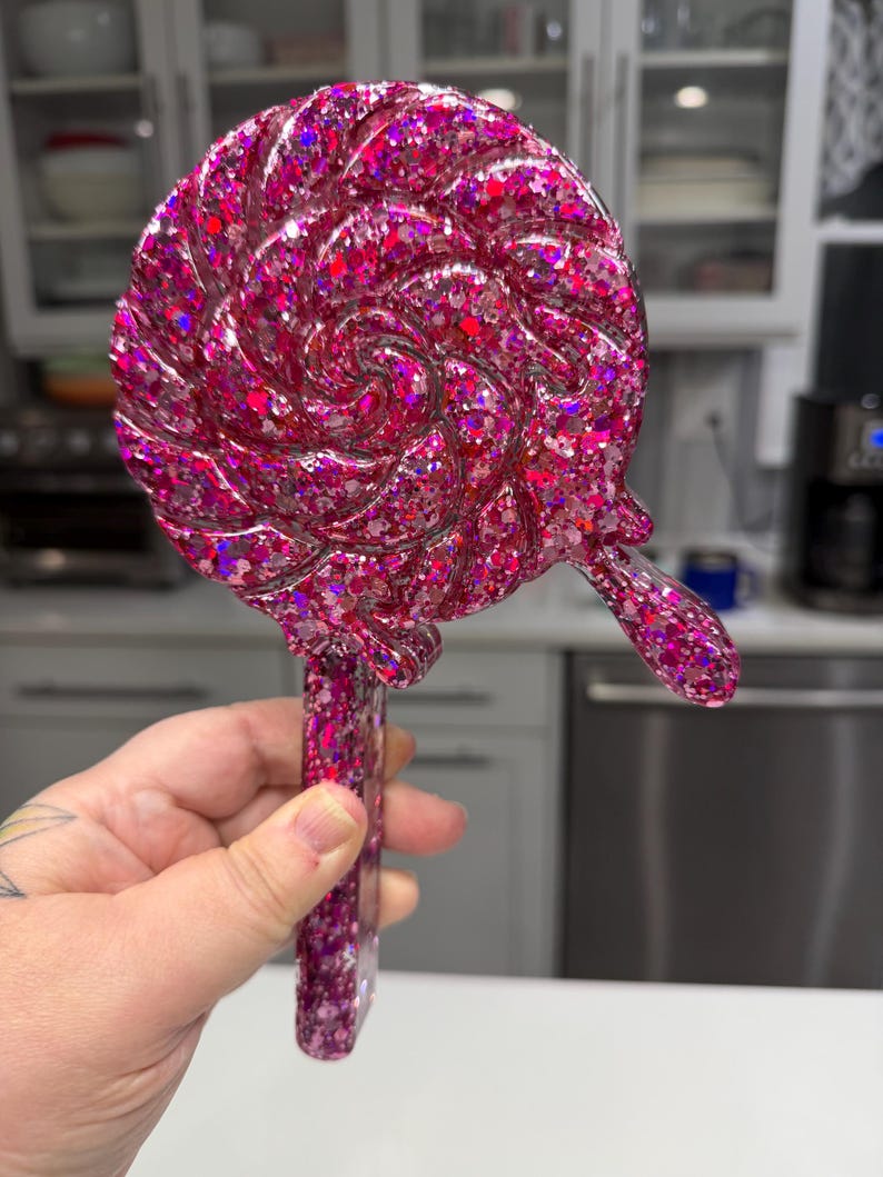 May include: A lollipop-shaped object featuring a swirling, circular design with a thick handle. The entire piece is crafted from a translucent, deep pink resin material embedded with a dense mixture of reflective, multi-sized pink glitter.