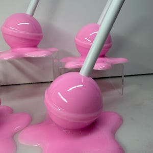 Op de afbeelding: Roze lolly-vormige decoraties met witte stokjes. De lolly's zijn rond en staan op een roze, gesmolten uitziende basis. Er zijn verschillende van deze decoratieve stukken te zien, wat wijst op een snoep- of dessertthema.