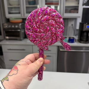 May include: A handheld mirror shaped like a round swirl lollipop, featuring a vibrant pink resin finish embedded with numerous reflective glitter particles. The circular mirror head includes a small decorative drip detail on the lower right side.