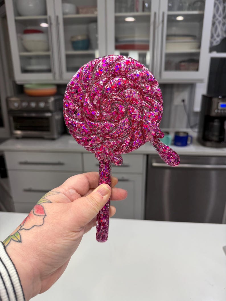 May include: A handheld mirror shaped like a round swirl lollipop, featuring a vibrant pink resin finish embedded with numerous reflective glitter particles. The circular mirror head includes a small decorative drip detail on the lower right side.