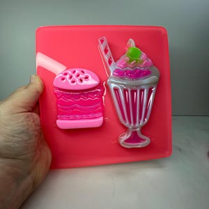 May include: A bright pink, rectangular resin piece featuring a burger and milkshake design. The burger has a dotted top bun, and the milkshake has a pink swirl and a green cherry on top. The design is in a cartoon style.