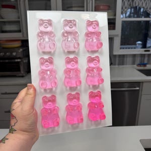 Pu&ograve; includere: Uno stampo di plastica trasparente riempito con dodici caramelle gommose a forma di orsetto rosa. Gli orsetti gommosi sono disposti in tre file da quattro. Le caramelle hanno un aspetto traslucido e sono di una tonalit&agrave; rosa vibrante. Lo stampo &egrave; tenuto su uno sfondo bianco.