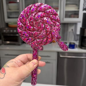 May include: A handheld mirror shaped like a round lollipop with a swirling pattern. The entire surface is covered in dense, vibrant pink and magenta glitter, featuring a long, straight handle and a small, decorative drip detail on the side.