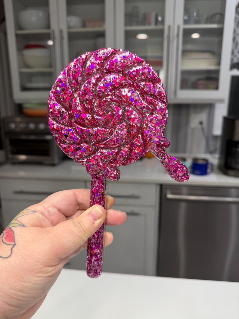 May include: A handheld mirror shaped like a round lollipop with a swirling pattern. The entire surface is covered in dense, vibrant pink and magenta glitter, featuring a long, straight handle and a small, decorative drip detail on the side.
