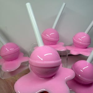 Op de afbeelding: Een verzameling roze lolly-vormige objecten met witte stokjes. Elke lolly staat op een roze, gesmolten uitziende basis. De objecten zijn gerangschikt op een helder oppervlak tegen een effen achtergrond. De lolly's hebben een glanzende afwerking.