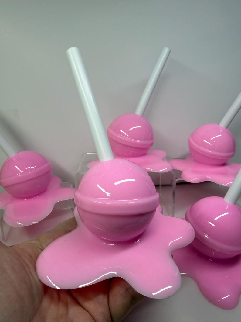 Op de afbeelding: Een verzameling roze lolly-vormige objecten met witte stokjes. Elke lolly staat op een roze, gesmolten uitziende basis. De objecten zijn gerangschikt op een helder oppervlak tegen een effen achtergrond. De lolly's hebben een glanzende afwerking.