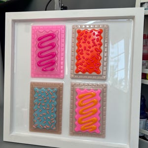 May include: A framed artwork featuring four colorful, stylized Pop-Tart designs. Each rectangular design has a different color frosting and sprinkles. The frame is white, and the background is a glossy white surface. The Pop-Tarts are pink, orange, blue, and pink with orange frosting.