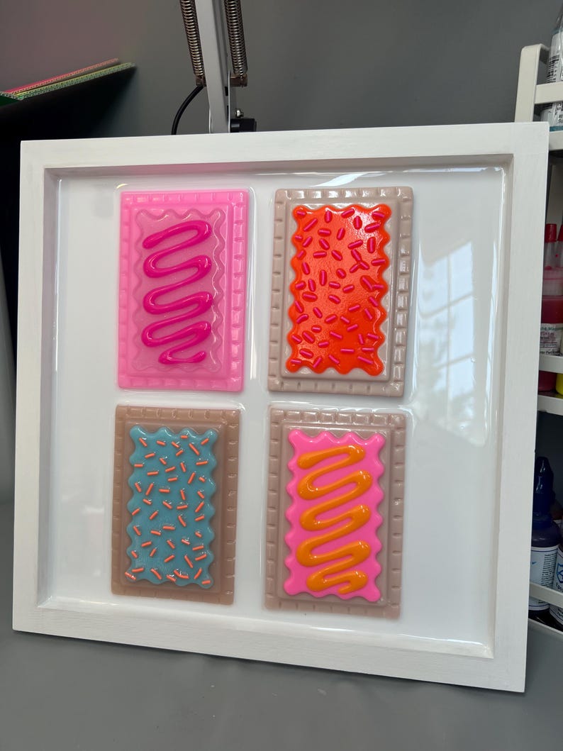 May include: A framed artwork featuring four colorful, stylized Pop-Tart designs. Each rectangular design has a different color frosting and sprinkles. The frame is white, and the background is a glossy white surface. The Pop-Tarts are pink, orange, blue, and pink with orange frosting.