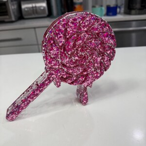 May include: A clear resin lollipop-shaped ornament filled with vibrant pink glitter. The circular head features a swirling pattern with a dripping effect, and the straight handle extends diagonally. The entire piece has a glossy finish.
