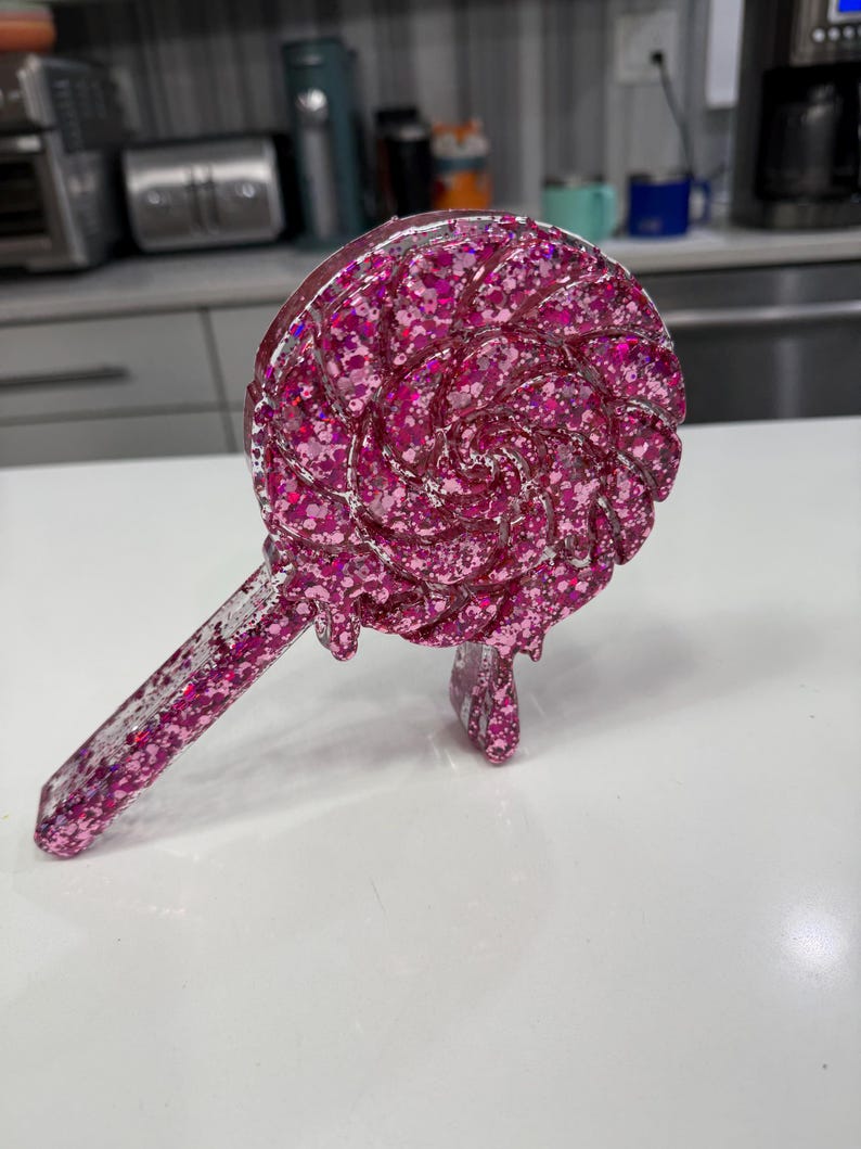 May include: A clear resin lollipop-shaped ornament filled with vibrant pink glitter. The circular head features a swirling pattern with a dripping effect, and the straight handle extends diagonally. The entire piece has a glossy finish.