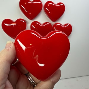 May include: A selection of glossy, red heart-shaped objects. One large heart is held in a hand, with several smaller hearts arranged around it. The hearts have a smooth, reflective surface, possibly made of glass or a similar material.