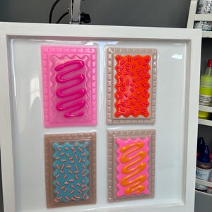 May include: A framed artwork featuring four colorful, rectangular designs resembling frosted pastries. Each pastry has a different color frosting and sprinkles, including pink, orange, blue, and yellow. The frame is white.
