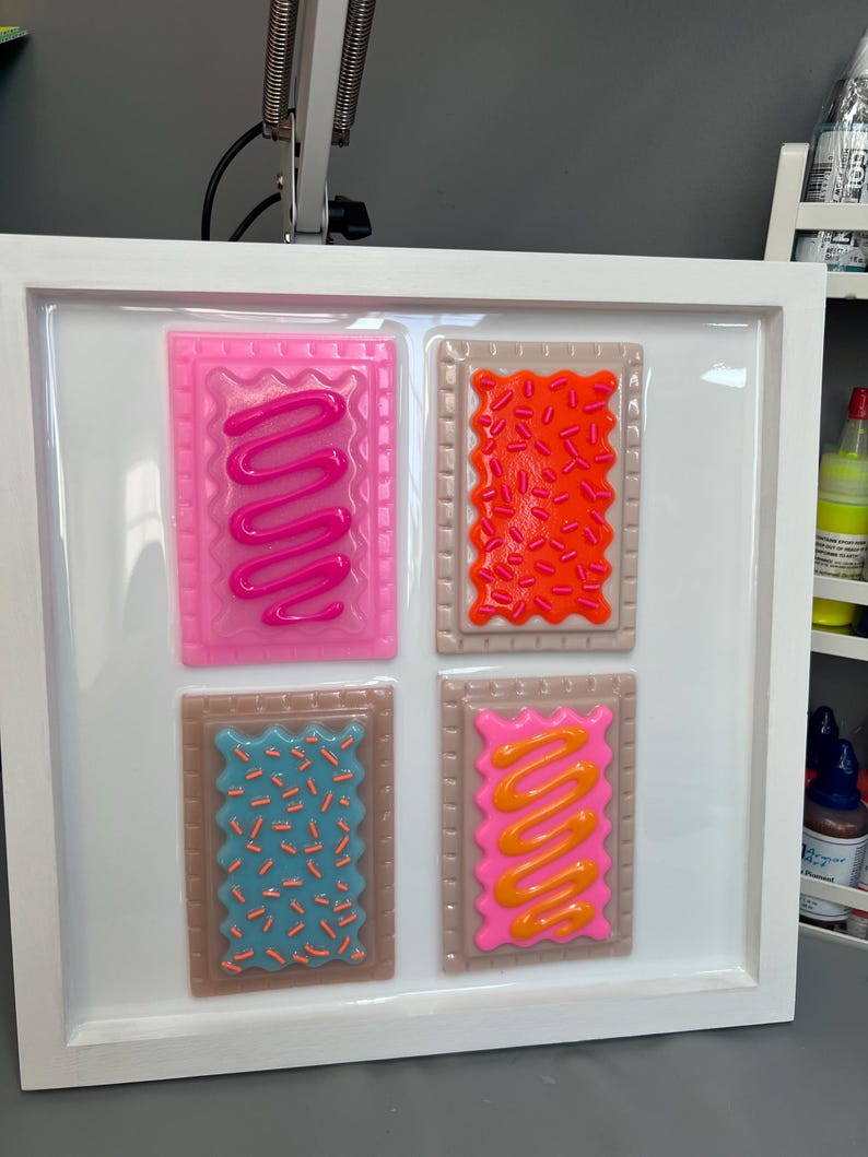 May include: A framed artwork featuring four colorful, rectangular designs resembling frosted pastries. Each pastry has a different color frosting and sprinkles, including pink, orange, blue, and yellow. The frame is white.