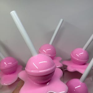 Op de afbeelding: Een verzameling roze lolly's met witte stokjes. Elke lolly heeft een ronde snoepkop en een gesmolten, druipende basis. De lolly's zijn glanzend en lijken van plastic of hars te zijn gemaakt.