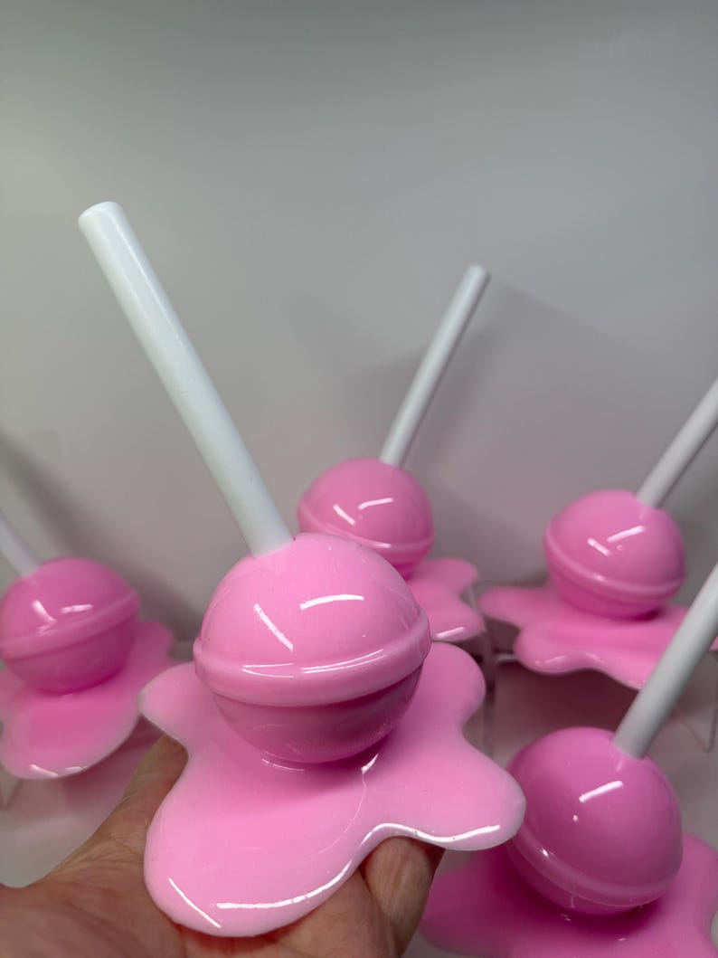 Op de afbeelding: Een verzameling roze lolly's met witte stokjes. Elke lolly heeft een ronde snoepkop en een gesmolten, druipende basis. De lolly's zijn glanzend en lijken van plastic of hars te zijn gemaakt.