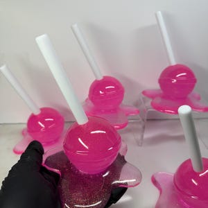 Puede incluir: Una colecci&oacute;n de piruletas rosas con purpurina y palitos blancos, sobre bases transparentes con aspecto de derretido. Las piruletas est&aacute;n dispuestas sobre una superficie blanca. Una de ellas es sostenida por una mano enguantada de negro. Aspecto brillante y transparente.