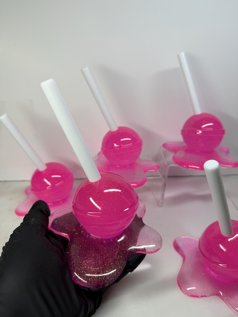 Puede incluir: Una colecci&oacute;n de piruletas rosas con purpurina y palitos blancos, sobre bases transparentes con aspecto de derretido. Las piruletas est&aacute;n dispuestas sobre una superficie blanca. Una de ellas es sostenida por una mano enguantada de negro. Aspecto brillante y transparente.