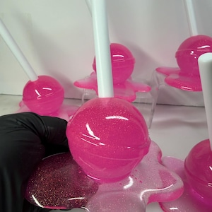 Puede incluir: Varias piruletas rosas con purpurina y palitos blancos. Las piruletas, de forma esf&eacute;rica, se asientan sobre una base rosa, brillante y transparente. La imagen destaca el color rosa vibrante y la textura brillante.