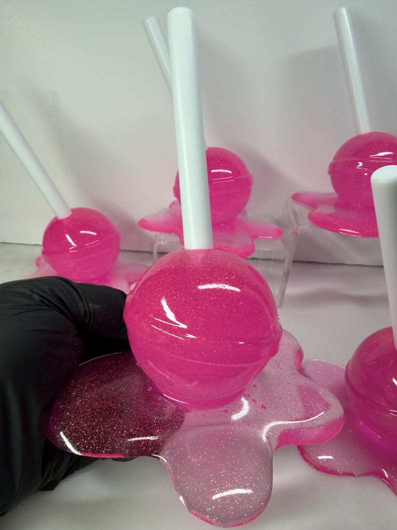 Puede incluir: Varias piruletas rosas con purpurina y palitos blancos. Las piruletas, de forma esf&eacute;rica, se asientan sobre una base rosa, brillante y transparente. La imagen destaca el color rosa vibrante y la textura brillante.