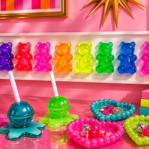 May include: A collection of colorful resin decor featuring a wall-mounted display of eight translucent gummy bears in a rainbow gradient, three lollipop-shaped containers with white straws, and two heart-shaped beaded trays holding small spheres.