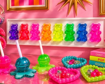 Puede incluir: Una colección de decoración de resina colorida que incluye un expositor de pared con ocho ositos de goma translúcidos en degradado arcoíris, tres recipientes con forma de piruleta con pajitas blancas y dos bandejas de cuentas en forma de corazón.