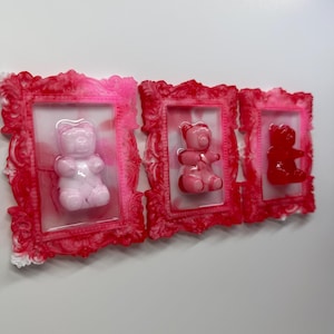 May include: Three framed resin art pieces featuring gummy bear sculptures. Each frame is a vibrant pink with ornate detailing. The gummy bears are in shades of pink, ranging from light to dark, creating a playful and colorful wall art display.