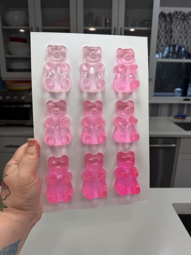 Pu&ograve; includere: Uno stampo di plastica trasparente riempito con dodici caramelle gommose a forma di orsetto rosa. Gli orsetti gommosi sono disposti in tre file da tre, con diverse sfumature di rosa. Lo stampo &egrave; tenuto in mano, con uno sfondo di cucina.