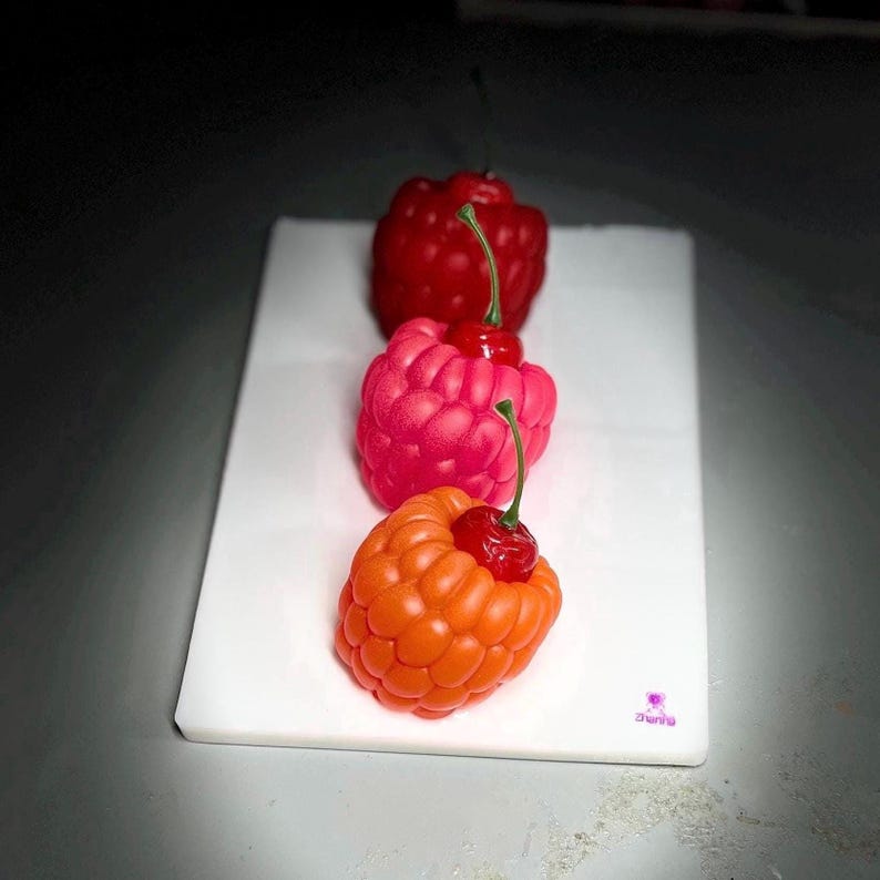 May include: Three colorful raspberry-shaped candles with cherry accents are arranged on a white rectangular surface. The candles are in shades of red, pink, and orange. The cherries have green stems.