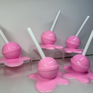 Op de afbeelding: Een verzameling roze lolly's met witte stokjes, die lijken te smelten. De lolly's zijn rond en glanzend, met het gesmolten snoepgoed dat een plasvorm vormt.