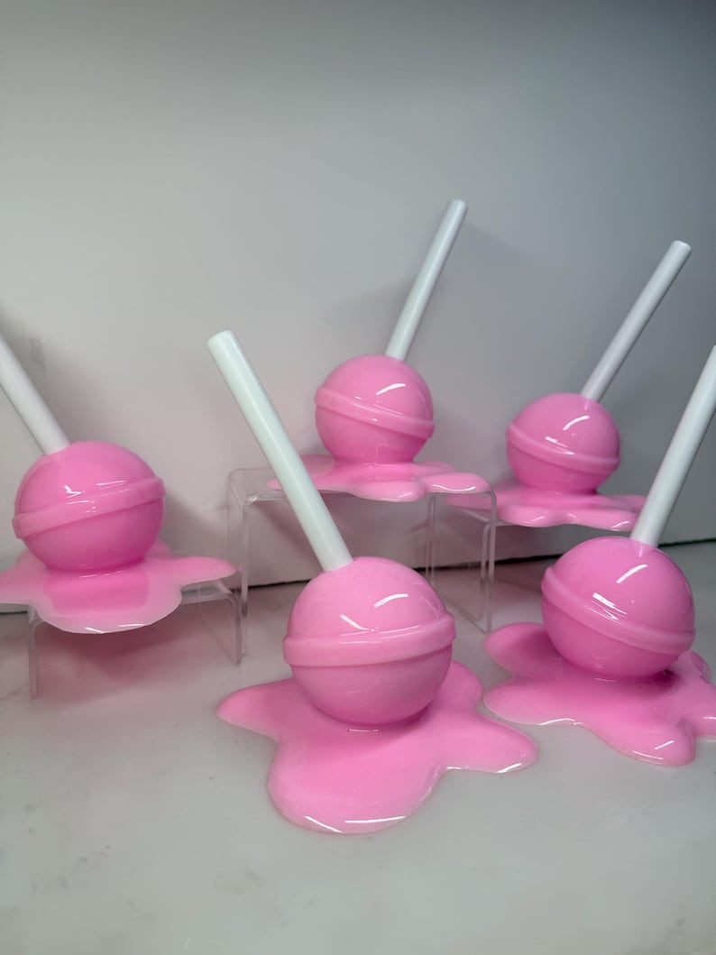Op de afbeelding: Een verzameling roze lolly's met witte stokjes, die lijken te smelten. De lolly's zijn rond en glanzend, met het gesmolten snoepgoed dat een plasvorm vormt.