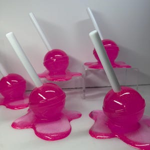 Puede incluir: Varias piruletas de color rosa brillante con palitos blancos est&aacute;n dispuestas sobre una superficie blanca. Las piruletas parecen estar derriti&eacute;ndose, con el caramelo rosa extendi&eacute;ndose alrededor de la base de cada una. El caramelo tiene un acabado brillante.