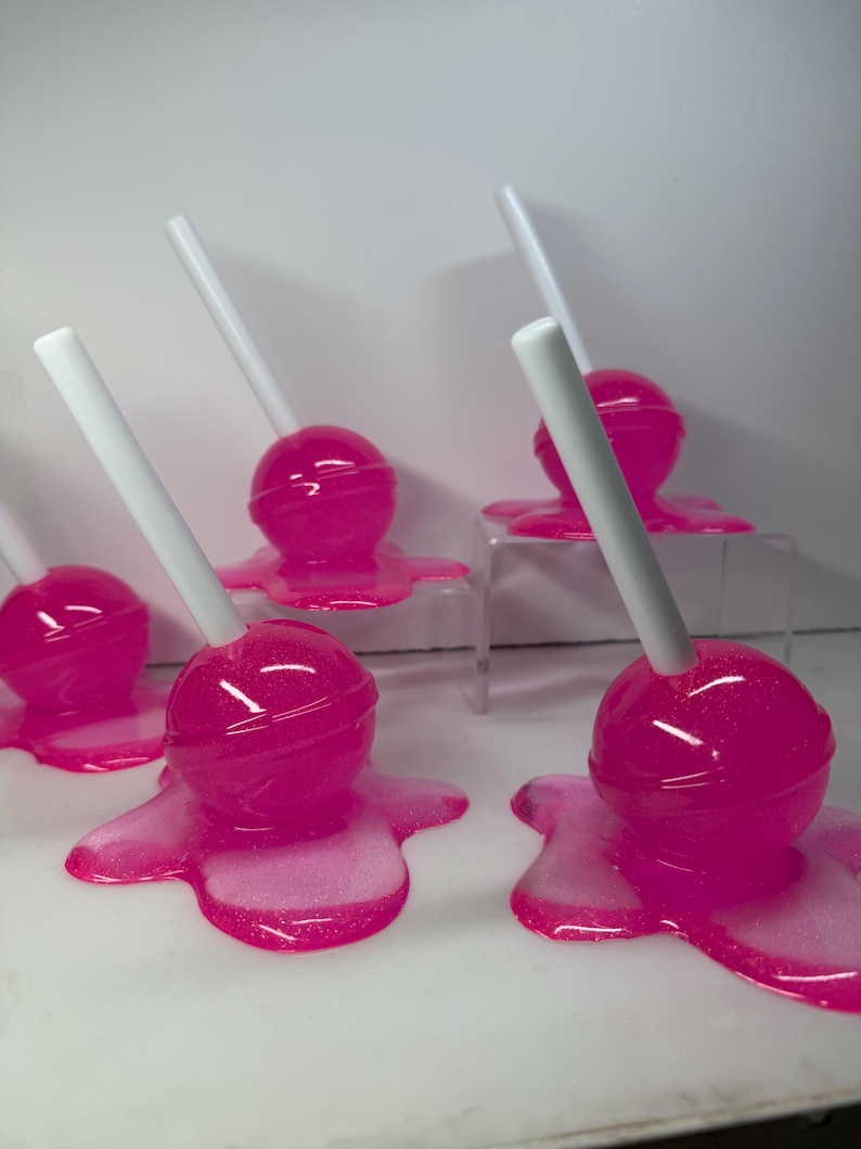Puede incluir: Varias piruletas de color rosa brillante con palitos blancos est&aacute;n dispuestas sobre una superficie blanca. Las piruletas parecen estar derriti&eacute;ndose, con el caramelo rosa extendi&eacute;ndose alrededor de la base de cada una. El caramelo tiene un acabado brillante.