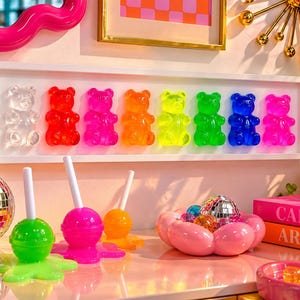 May include: A white rectangular wall frame containing eight translucent gummy bear figures in clear, red, pink, orange, yellow, green, blue, and purple. Below are three lollipop sculptures in green, pink, and orange, and a pink flower-shaped bowl.