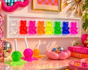 Puede incluir: Un marco de pared rectangular blanco que contiene ocho figuras de ositos de goma translúcidos en transparente, rojo, rosa, naranja, amarillo, verde, azul y morado. Debajo hay tres esculturas de piruletas en verde, rosa y naranja y un cuenco rosa.