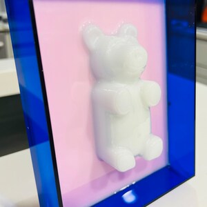 Puede incluir: Una pieza de arte mural rectangular que presenta una figura de oso de goma tridimensional de color blanco brillante centrada sobre un fondo rosa claro plano, todo encerrado en un marco de acr&iacute;lico azul real transl&uacute;cido.