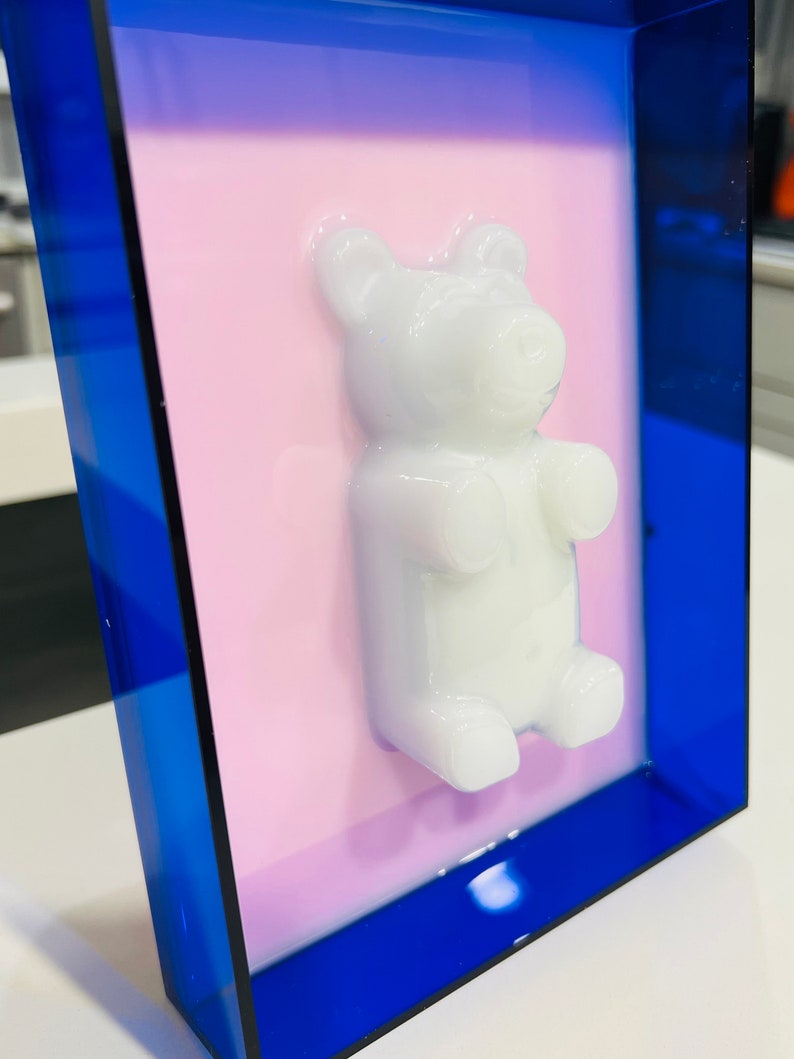 Puede incluir: Una pieza de arte mural rectangular que presenta una figura de oso de goma tridimensional de color blanco brillante centrada sobre un fondo rosa claro plano, todo encerrado en un marco de acr&iacute;lico azul real transl&uacute;cido.