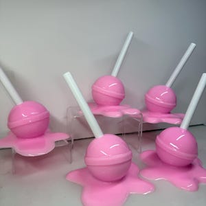 Op de afbeelding: Roze lolly's met witte stokjes, die lijken te smelten op een bijpassend roze oppervlak. De lolly's zijn rond met een ring in het midden. De afbeelding toont meerdere objecten met een snoepthema.