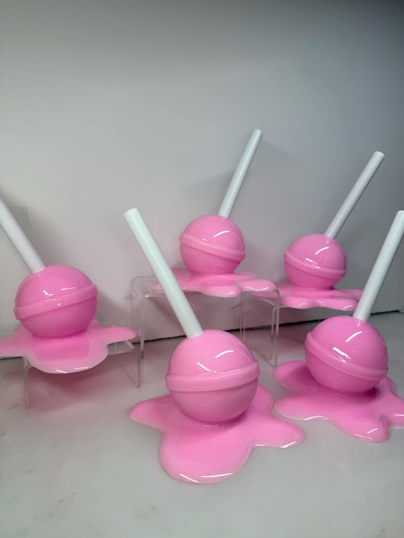 Op de afbeelding: Roze lolly's met witte stokjes, die lijken te smelten op een bijpassend roze oppervlak. De lolly's zijn rond met een ring in het midden. De afbeelding toont meerdere objecten met een snoepthema.